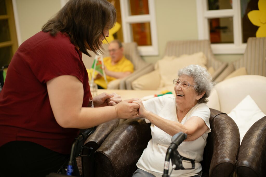 A woman standing up helps an older woman up from a chair. They are both smiling.